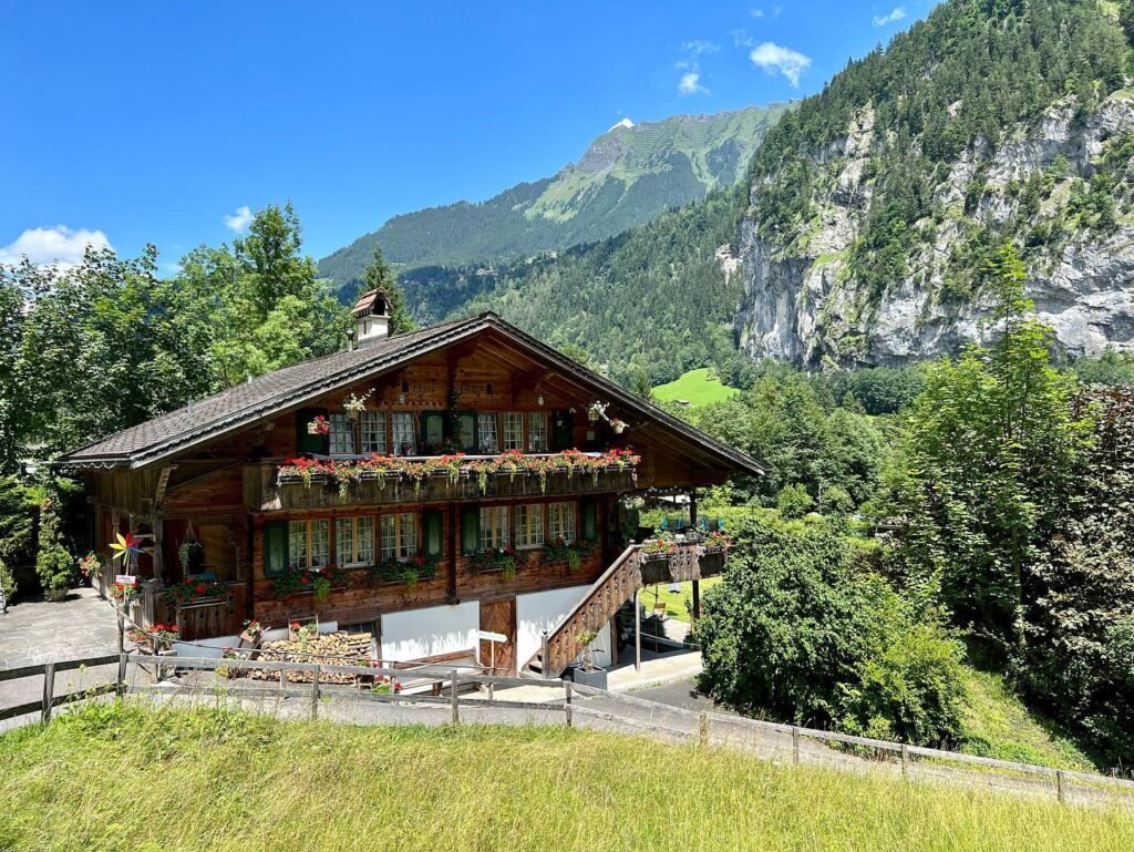 Traditional wooden chalets with flower-filled balconies nestled in lush green grasslands beneath towering rugged cliffs and a bright blue alpine sky.