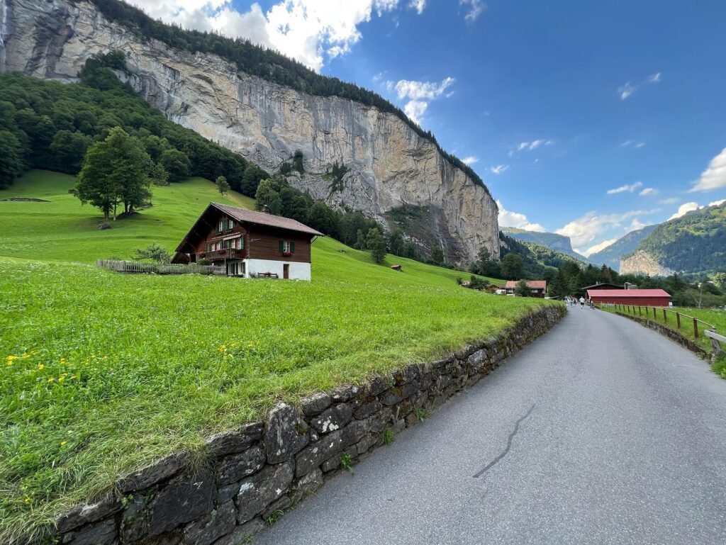 A mountain road runs past a traditional wooden chalet and green pasture beneath a cliff face.