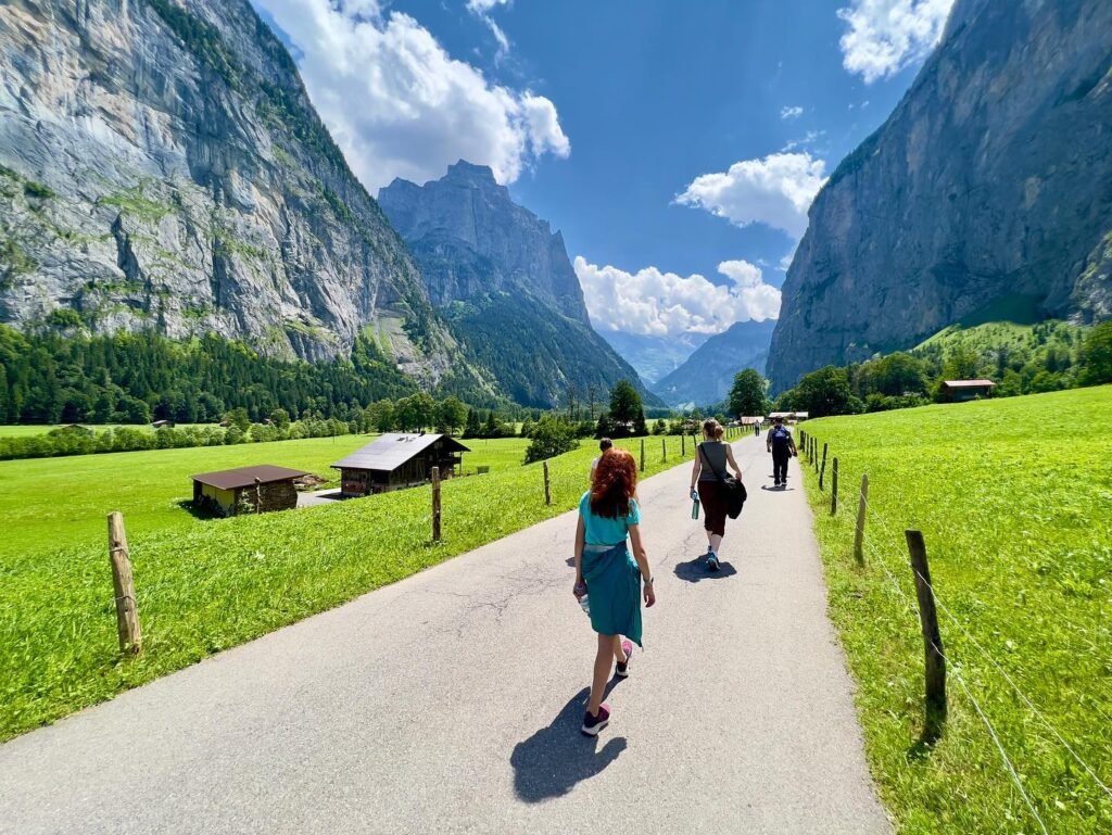 People walking a trail in a lush green valley with steep granite cliffs towering overhead. Bright blue skies with clouds provide a stunning backdrop.