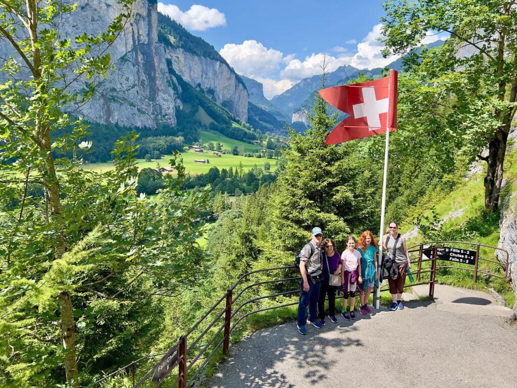 A scenic view of jagged Swiss mountains with lush greenery and a prominent Swiss flag at a paved trail overlook.
