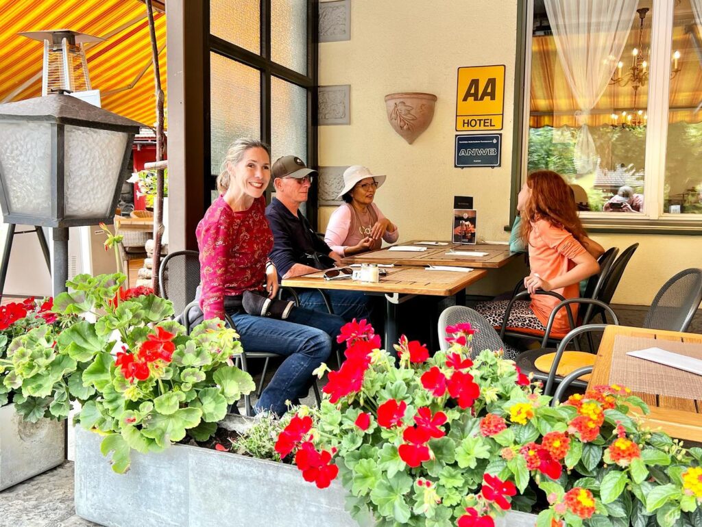 A group of people enjoy a sunny day at an outdoor café, with vibrant red and yellow flowers in the foreground and an AA hotel sign on the wall.