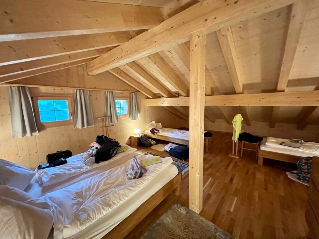 Interior of a rustic wooden cabin guestroom featuring multiple beds, exposed ceiling beams, and hard wood floors.