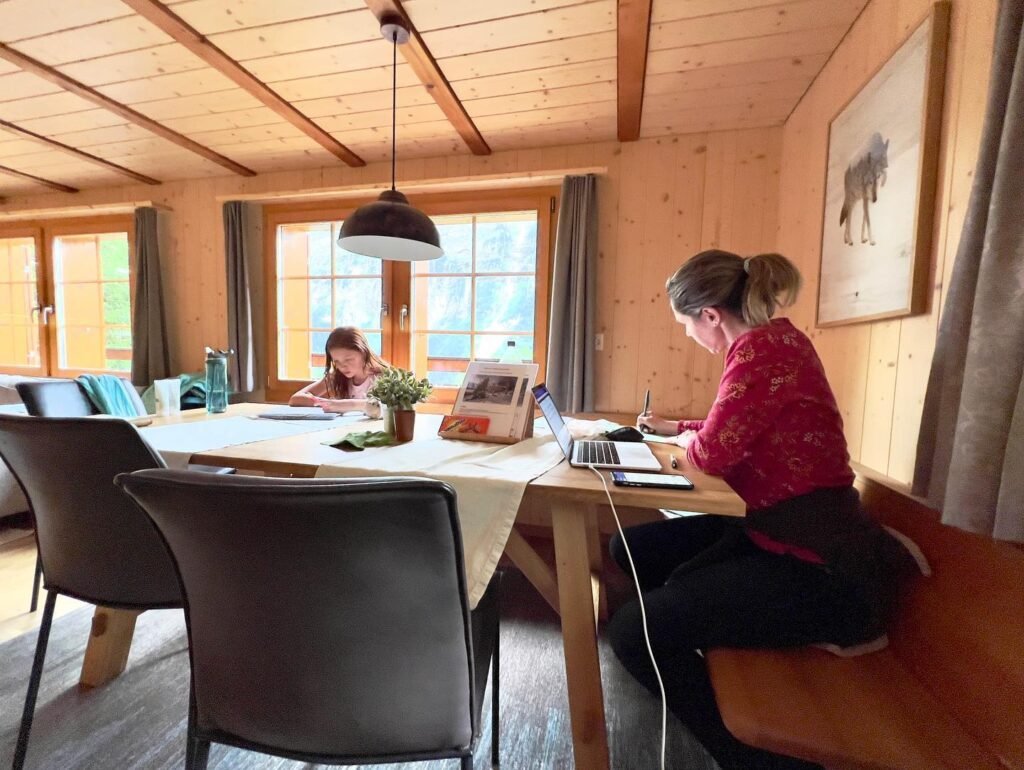 A cozy wooden interior with people working at a long table with laptops. The space features warm timber walls, large windows, and rustic decor.