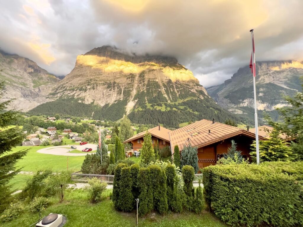 A wooden chalet with a red-tiled roof sits in a green landscape, with towering mountains illuminated by the sun behind it.