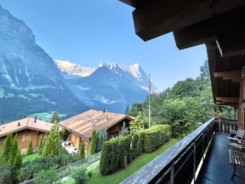A wooden balcony overlooking a picturesque mountain village with Alpine architecture and snow-capped peaks.