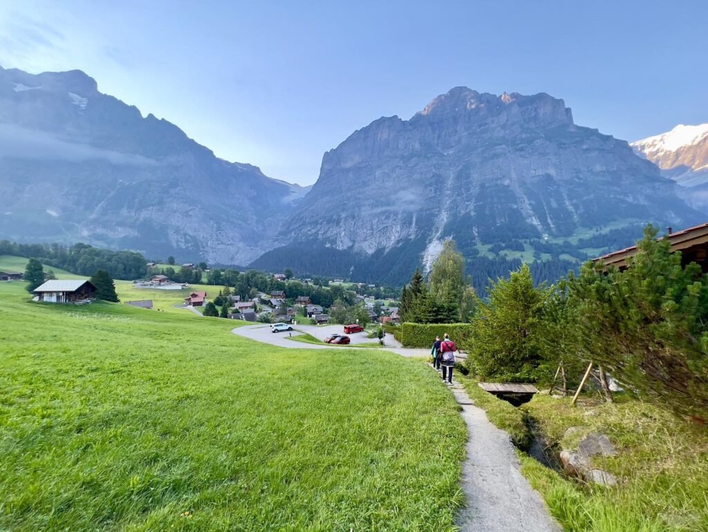 A gravel path winds through green fields with hikers heading toward distant mountains.