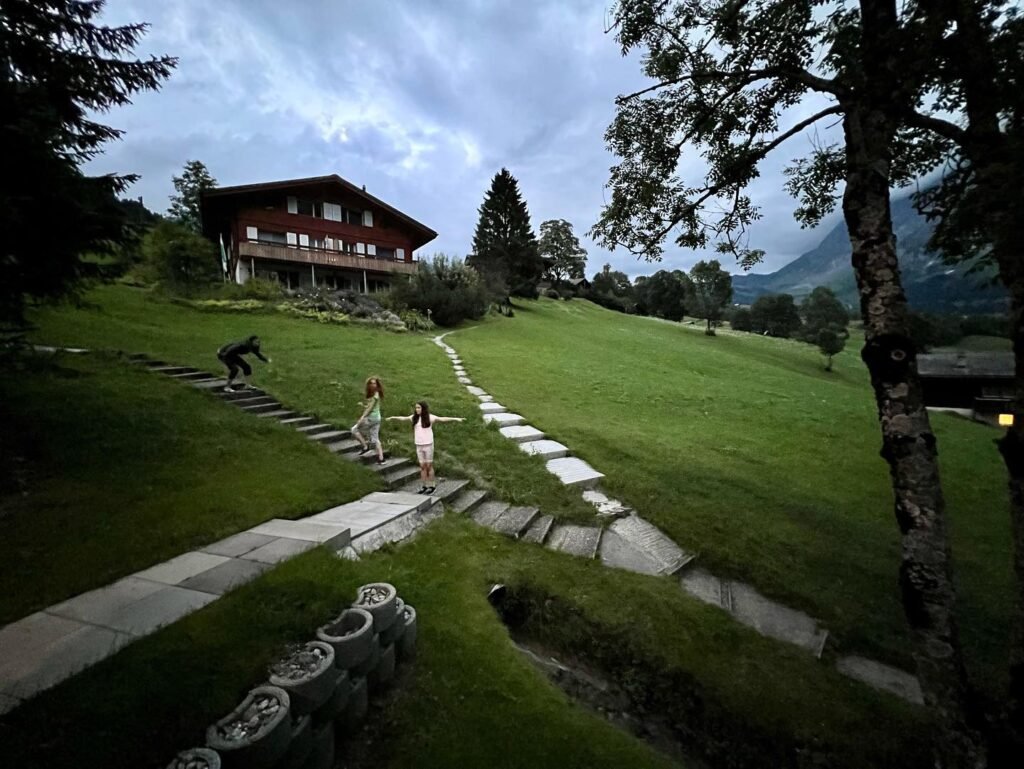 A grassy hillside leading to a wooden lodge with stone pathways under a cloudy sky. Several individuals walk on the path in the garden.