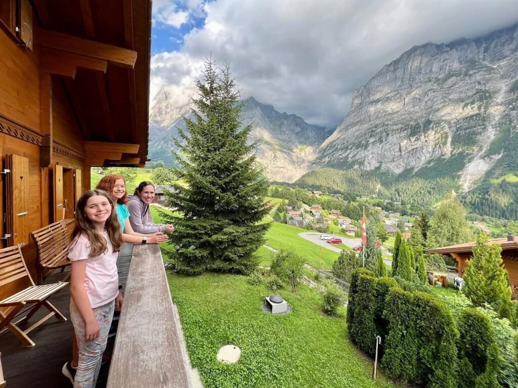 Family enjoying a scenic mountain view from a wooden balcony with lush greenery below. Dramatic skies overlook a picturesque valley and towering peaks.