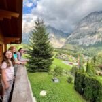 Schilthorn Family enjoying a scenic mountain view from a wooden balcony with lush greenery below. Dramatic skies overlook a picturesque valley and towering peaks.