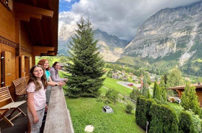 Family enjoying a scenic mountain view from a wooden balcony with lush greenery below. Dramatic skies overlook a picturesque valley and towering peaks.