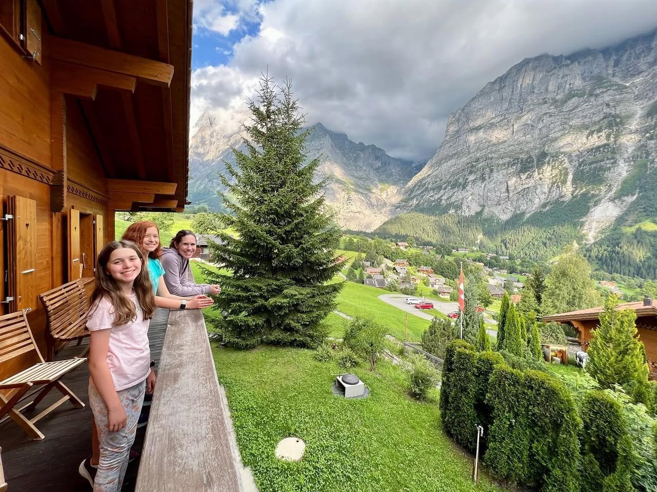 Grindelwald, Switzerland Family enjoying a scenic mountain view from a wooden balcony with lush greenery below. Dramatic skies overlook a picturesque valley and towering peaks.