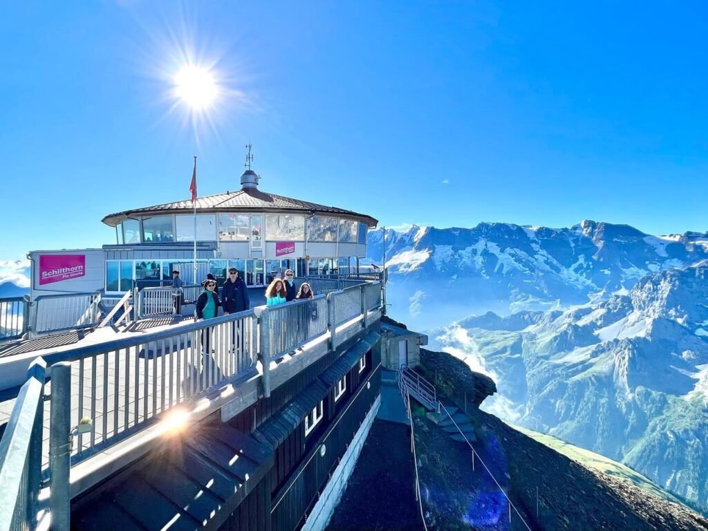 A scenic overlook at a mountain station offers stunning panoramic views of snow-capped peaks. The bright sun illuminates the clear blue sky.