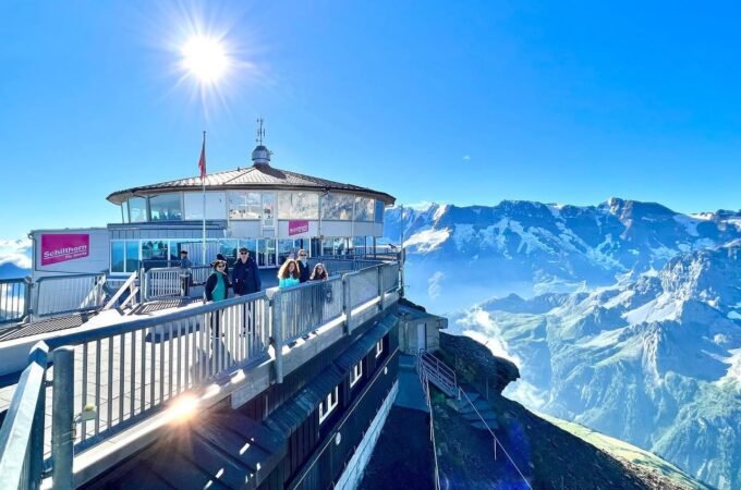 A scenic overlook at a mountain station offers stunning panoramic views of snow-capped peaks. The bright sun illuminates the clear blue sky.