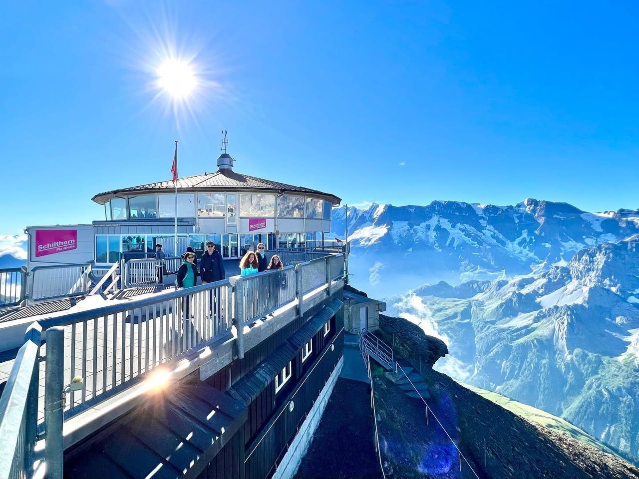 Schilthorn A scenic overlook at a mountain station offers stunning panoramic views of snow-capped peaks. The bright sun illuminates the clear blue sky.
