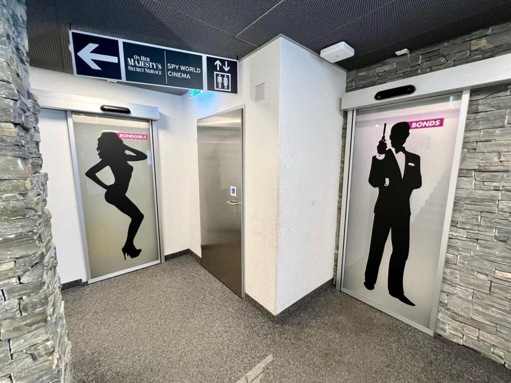 A modern airport restroom entrance features signage with directional arrows and large black silhouettes on white doors framed by stone walls.