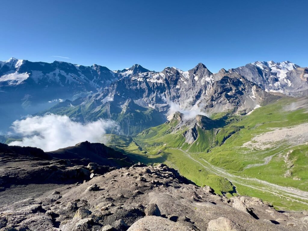Scenic mountain landscape showing granite peaks, a green alpine valley, and a distant trail beneath a clear blue sky.