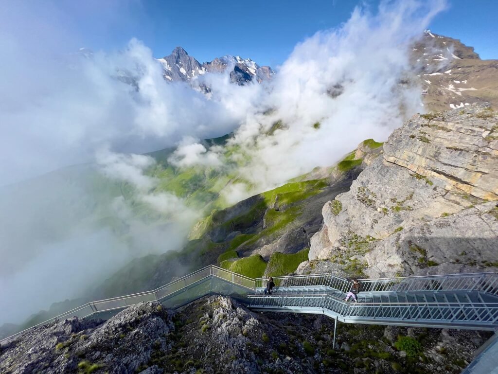 Workers construct a steel skybridge on a mountain peak with dramatic swirling clouds.