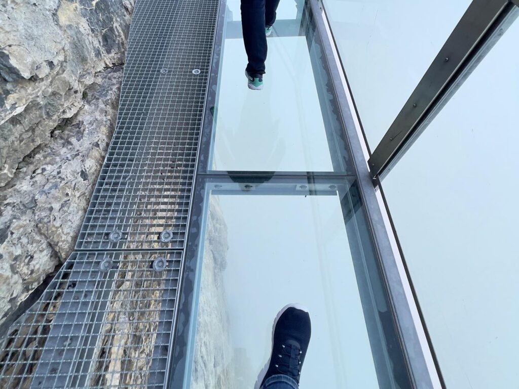 A person steps onto a transparent glass bridge with metal grating on one side. The view from below shows the rocky cliff face obscured by fog.