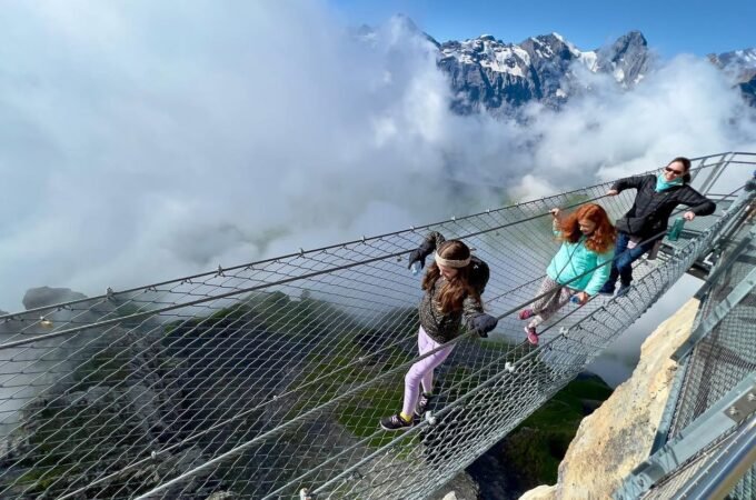 A group of people carefully crossing a suspended wire mesh bridge over a steep cliff with clouds in the background.