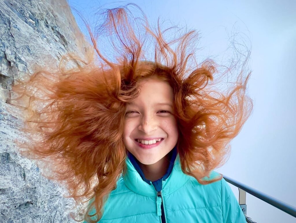 A young girl with red hair blowing in the wind smiles brightly while wearing a teal vest.