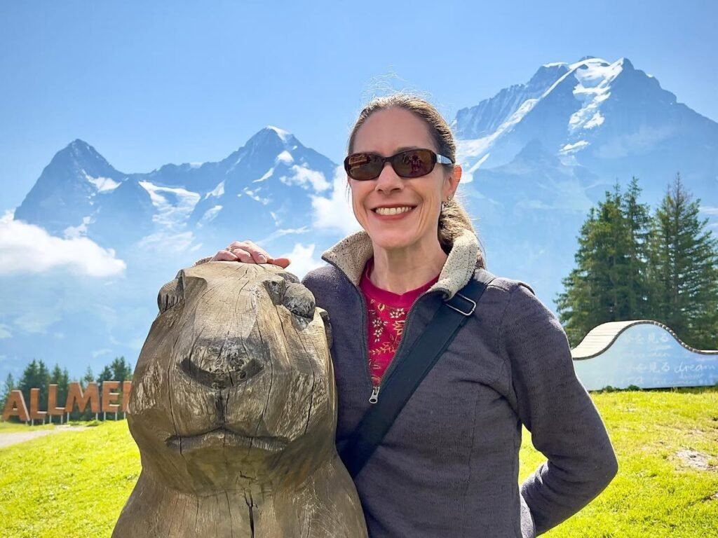 A traveler poses with a carved marmot statue at the Allmensattel welcome sign, set against a backdrop of snow-capped alpine peaks and green meadows.