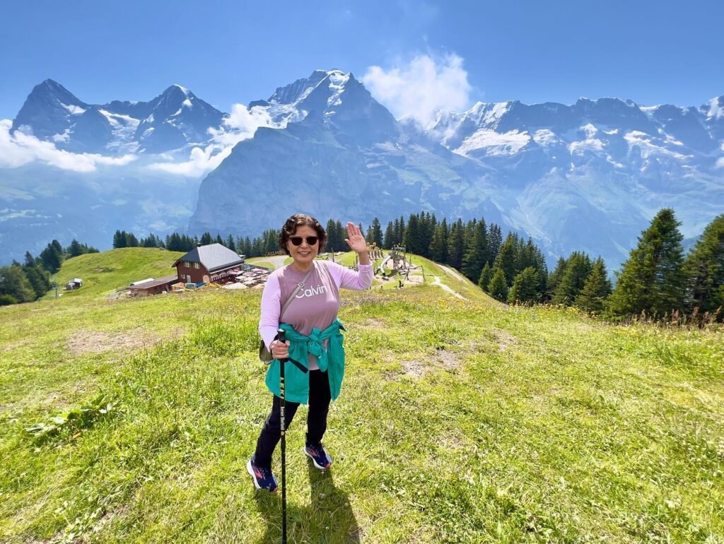 A hiker waves from a green meadow with snow-capped peaks and a mountain lake in the background.