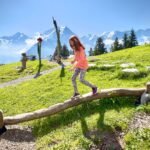 Skyline Walk A child in a pink shirt balances on a rustic log in a lush green alpine meadow, with snow-capped mountains in the distance.