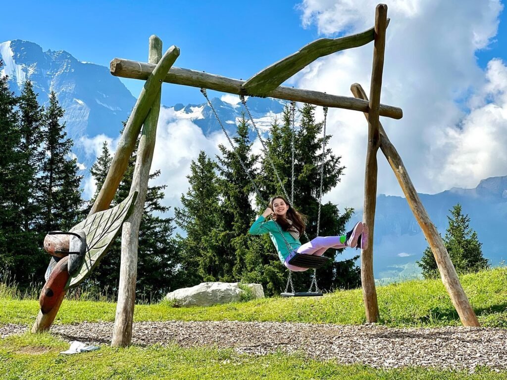 A person swings on a rustic wooden swing set against a backdrop of towering mountains and a bright blue sky.