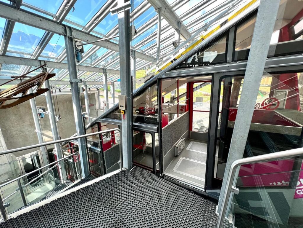 Interior of a cable car station with a glass roof and metal frames. The platform has a textured surface with railings.