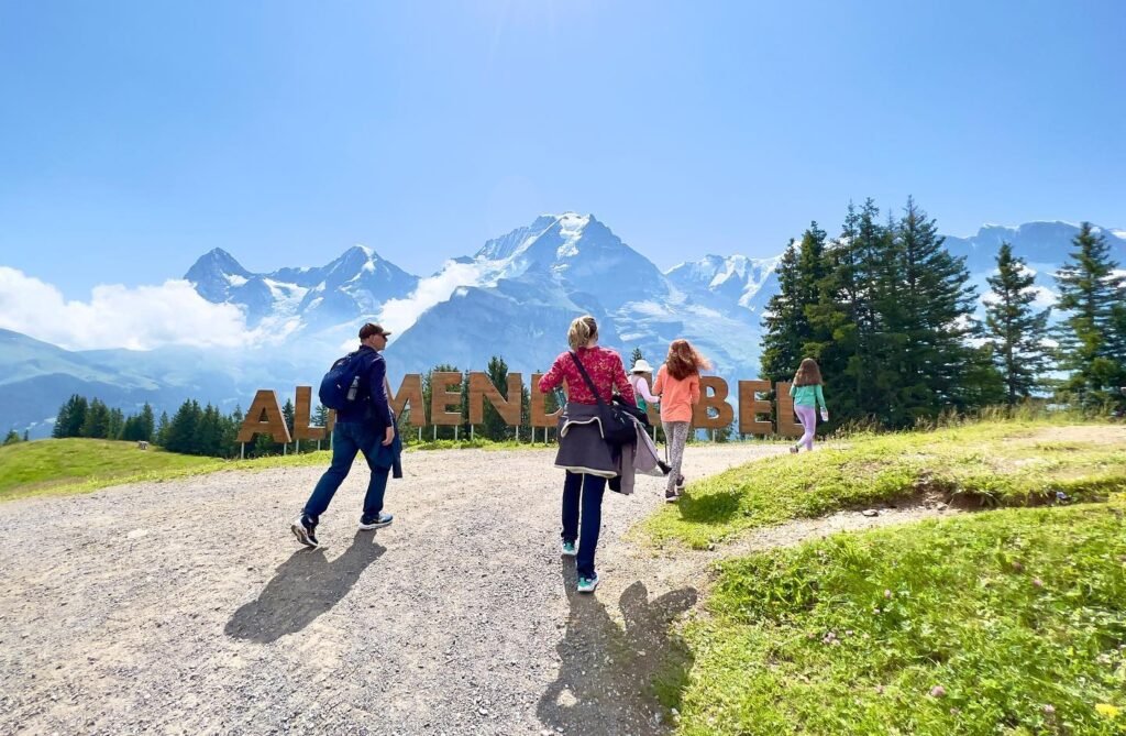 Visitors walking on a gravel path near the large 'Alpine Meadows' sign with snow-capped mountains and evergreen trees in the background.