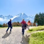 First Visitors walking on a gravel path near the large 'Alpine Meadows' sign with snow-capped mountains and evergreen trees in the background.