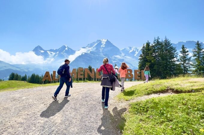 Visitors walking on a gravel path near the large 'Alpine Meadows' sign with snow-capped mountains and evergreen trees in the background.