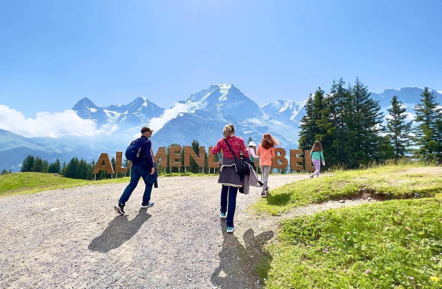 Mürren Visitors walking on a gravel path near the large 'Alpine Meadows' sign with snow-capped mountains and evergreen trees in the background.
