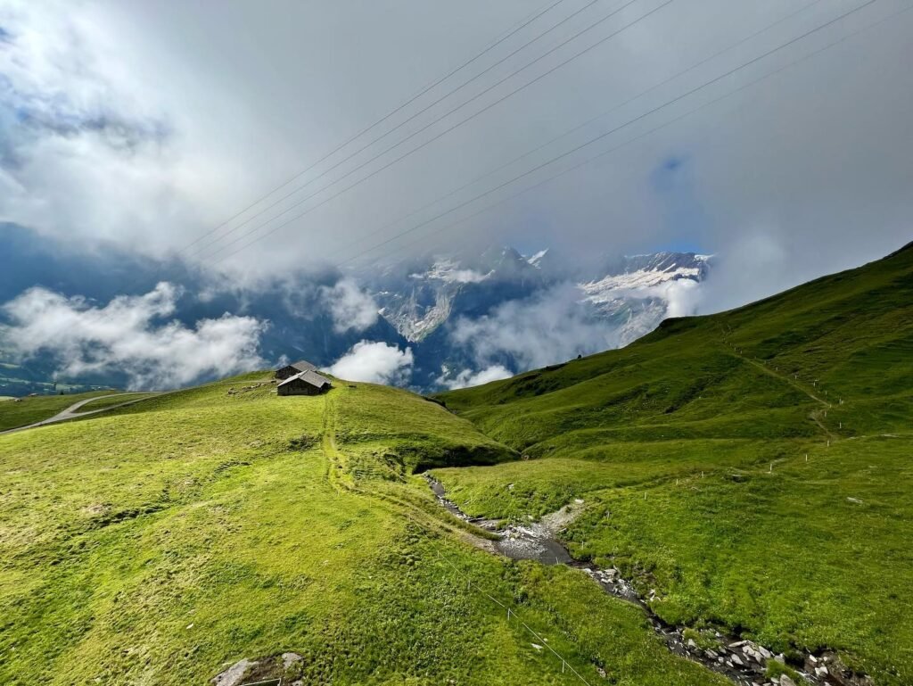 A scenic view of a mountain valley with a small wooden hut on a grassy ridge. The landscape features rolling green hills and a cloudy blue sky.
