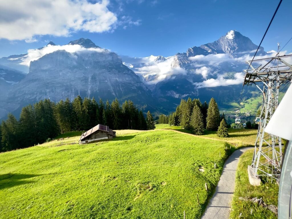 A scenic view from a cable car in the Alps, featuring vibrant green alpine meadows, a historic wooden hut, and towering snow-capped peaks. The mountains of Zermatt, including the iconic Matterhorn, create a striking contrast against the clear blue sky.