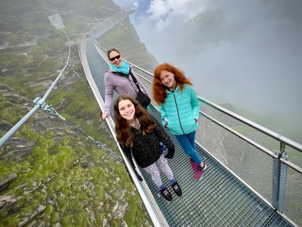 Three travelers on a glass-bottomed bridge in the mountains under a cloudy sky.