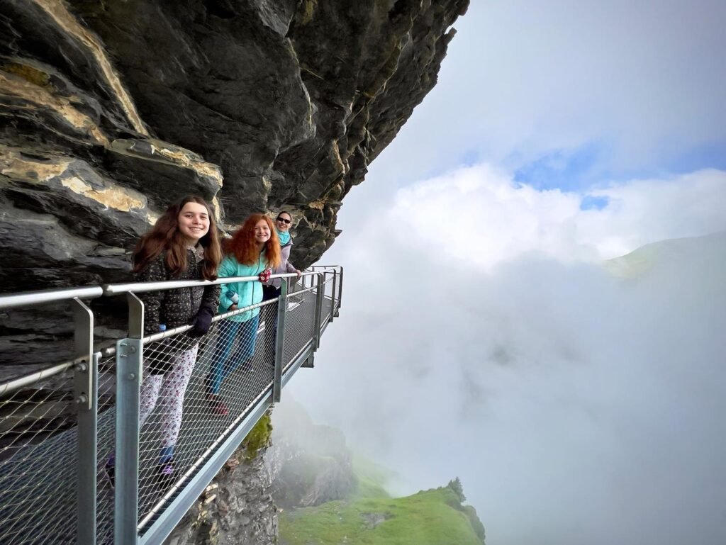 Two people walking along a misty cliffside path with dramatic rock formations and a view into the clouds.