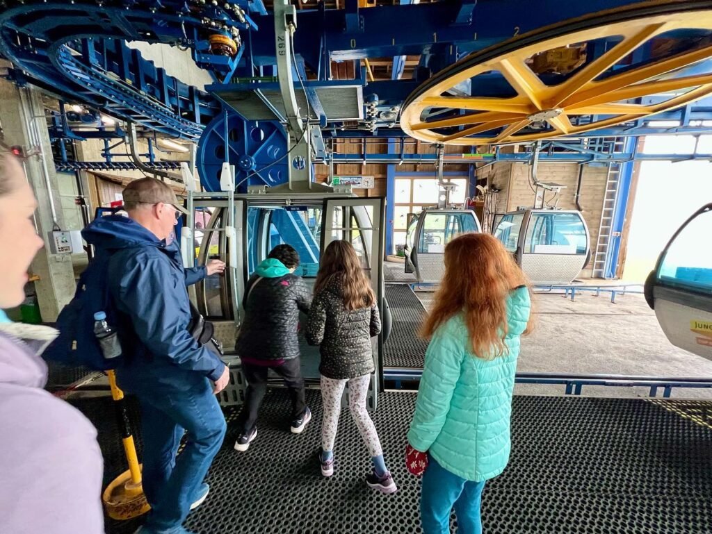 Visitors board and watch a traditional cable car at a station platform. Blue and yellow mechanical details are visible overhead.