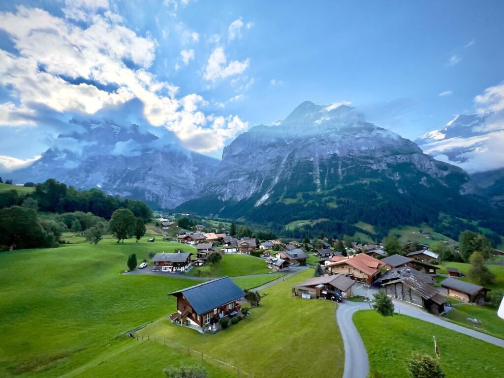 Aerial view of a peaceful alpine village with wooden homes set against a backdrop of dramatic mountains and a vibrant blue sky.
