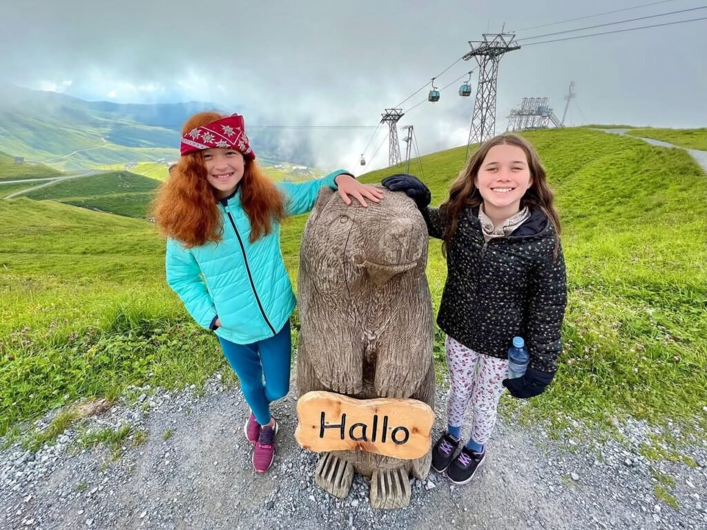 Two people posing with a moai statue on a scenic hillside with cable cars in the background under a cloudy sky. The setting includes a rocky path and rolling green terrain.