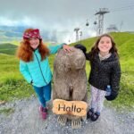 Mürren Two people posing with a moai statue on a scenic hillside with cable cars in the background under a cloudy sky. The setting includes a rocky path and rolling green terrain.