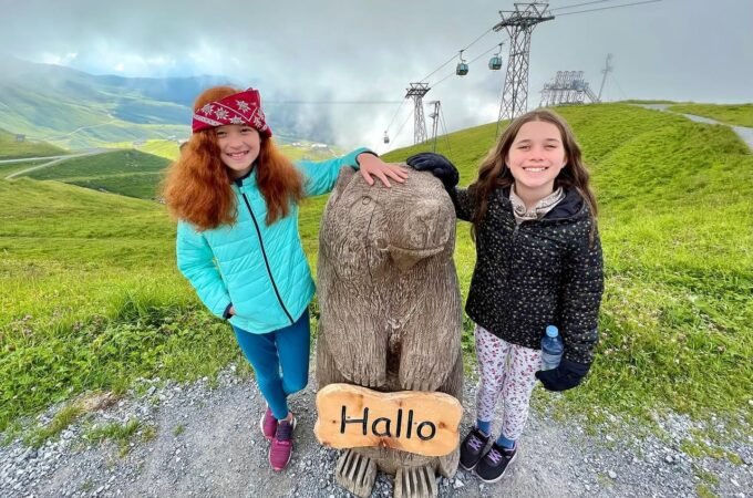 Two people posing with a moai statue on a scenic hillside with cable cars in the background under a cloudy sky. The setting includes a rocky path and rolling green terrain.