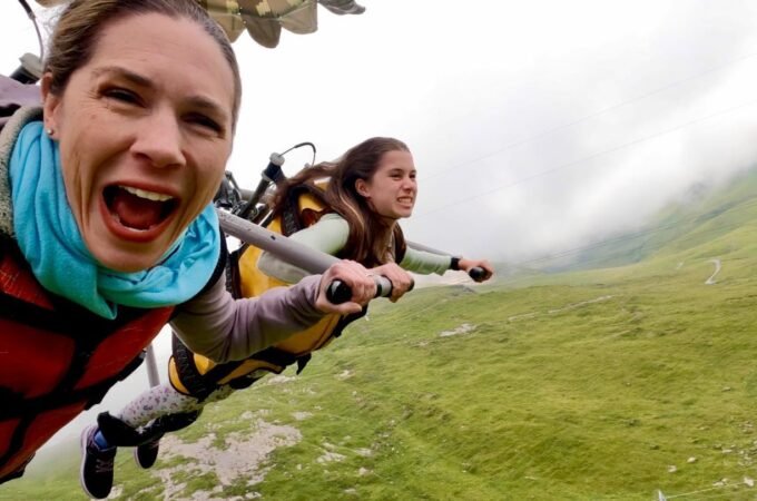 Two people in wingsuits glide over misty green hills in Scotland. The front person smiles with excitement while holding the control bar.