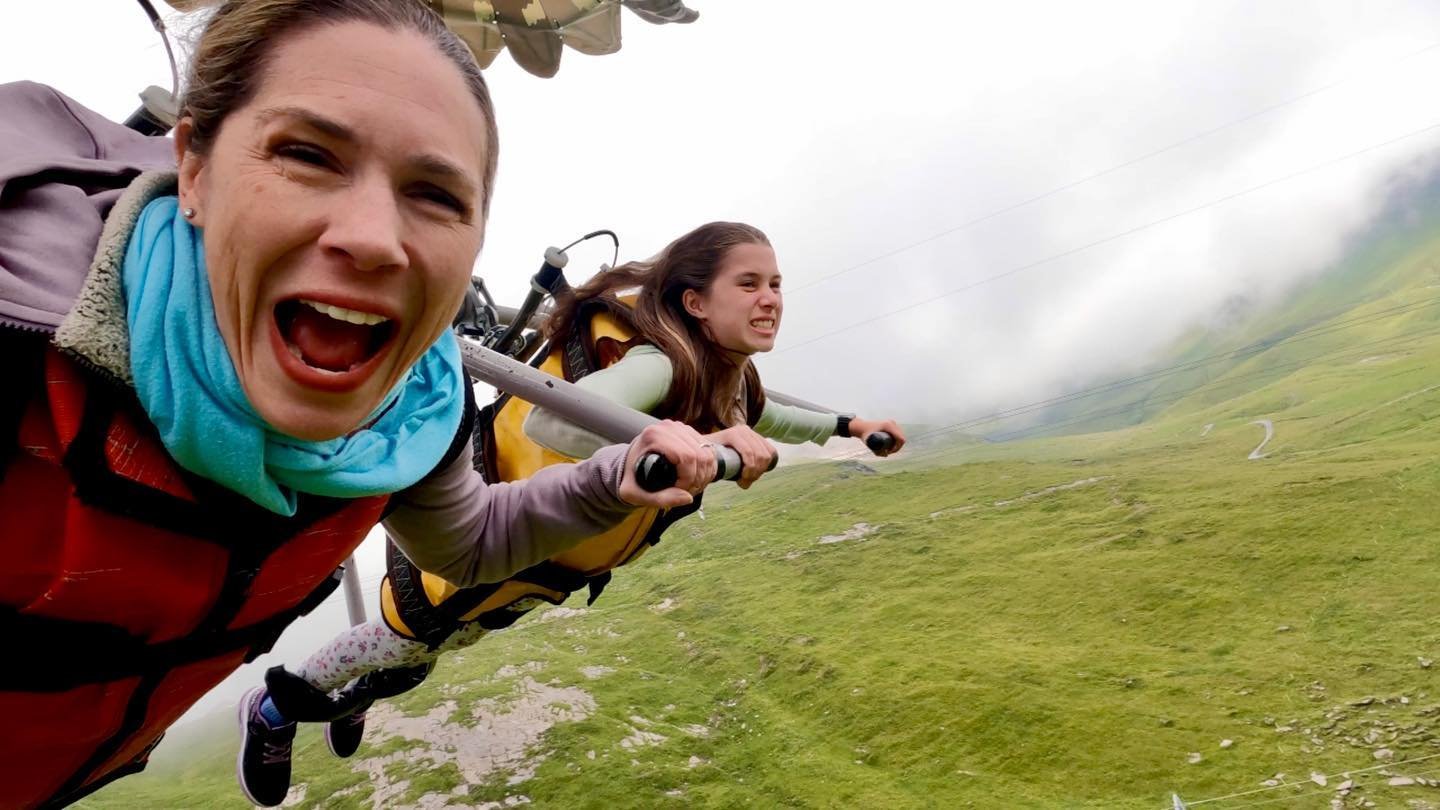 First Glider Two people in wingsuits glide over misty green hills in Scotland. The front person smiles with excitement while holding the control bar.