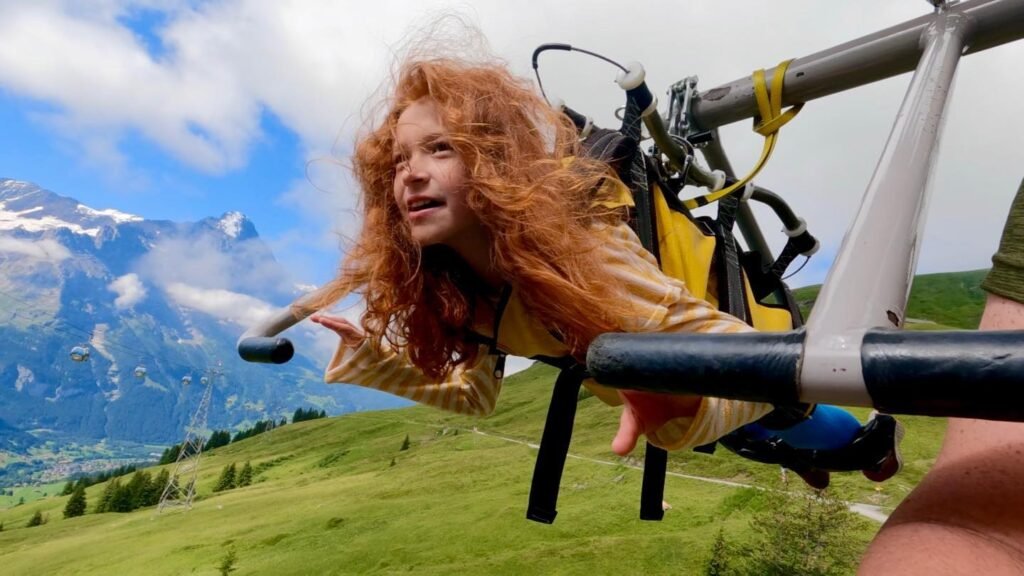 A person enjoys a thrilling ride on a swing suspended in a mountainous landscape with snowy peaks and green hills.