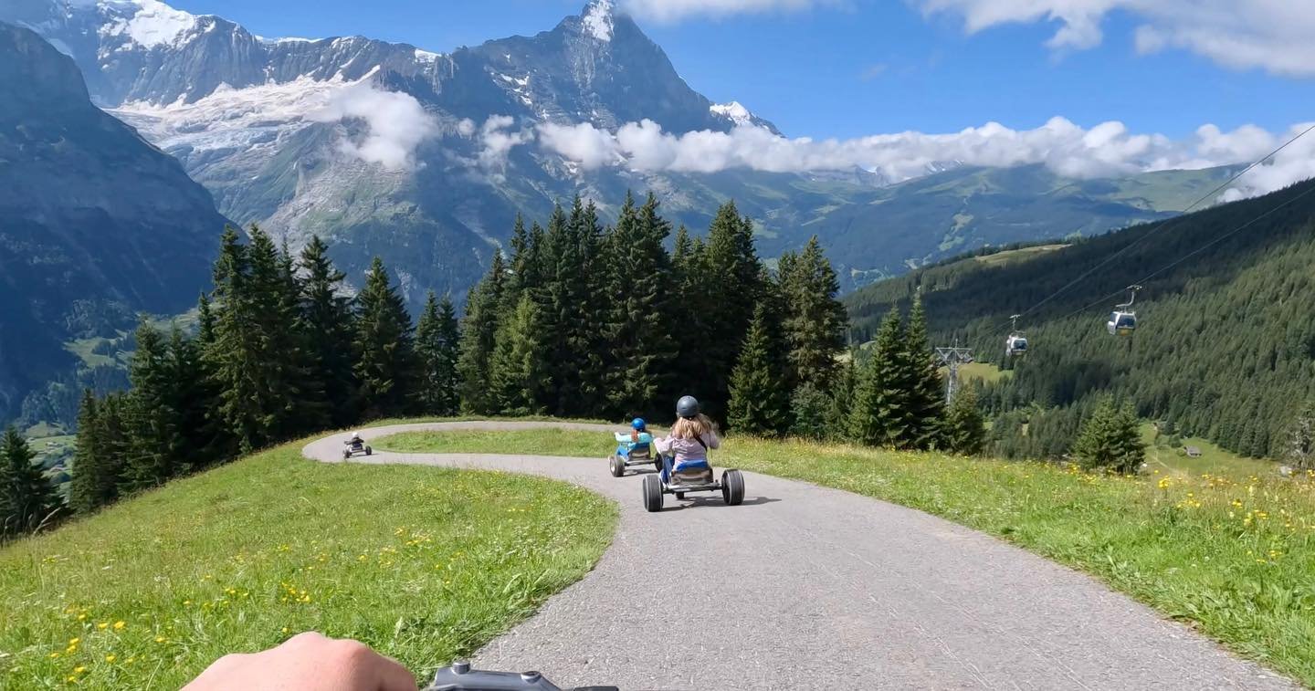 A paved path winds through lush green meadows with snow-capped mountains and drifts of clouds in the background. People are seen strolling along the path enjoying the scenic mountain vista.