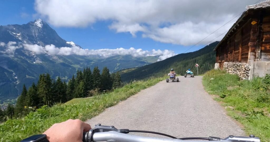 A view from a mountain bike riding along a scenic alpine road with snow-capped peaks and a rustic wooden cabin.