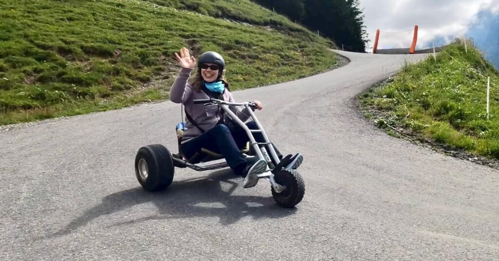 Pedaling a go-kart on a winding mountain road while waving at the camera.