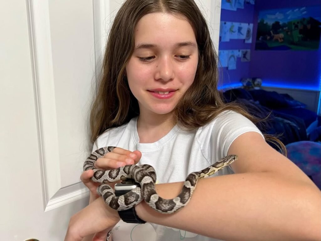 A young person gently holding a snake wrapped around their arm in a bedroom setting.