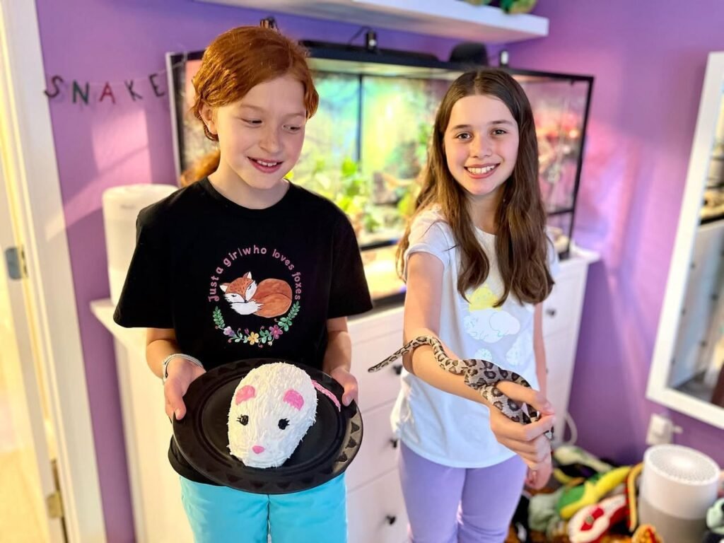 Children holding a small pet and a snake in a room with a reptile tank and the word 'SNAKE' on the wall.